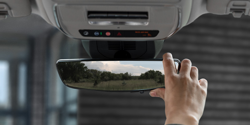 A hand adjusting the rearview mirror in a 2025 GMC Terrain Elevation, showing trees and field in the reflection.