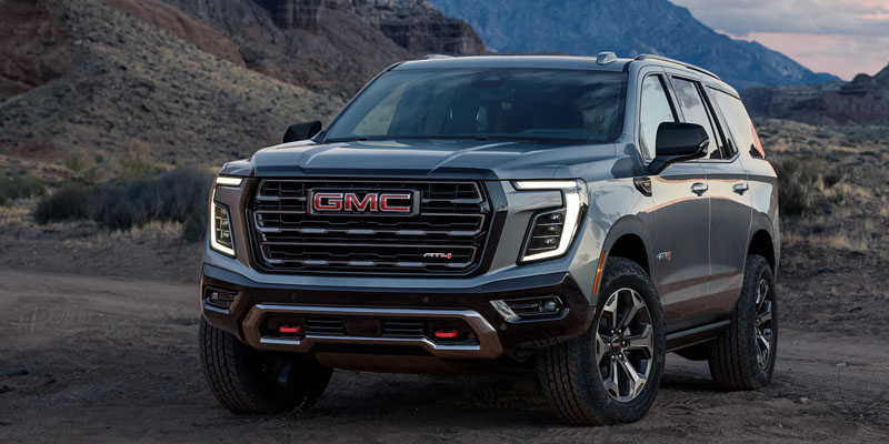 A rugged GMC Yukon XL parked on a dirt road, set against a mountainous backdrop at dusk. The vehicle's bold grille and sleek design convey strength.